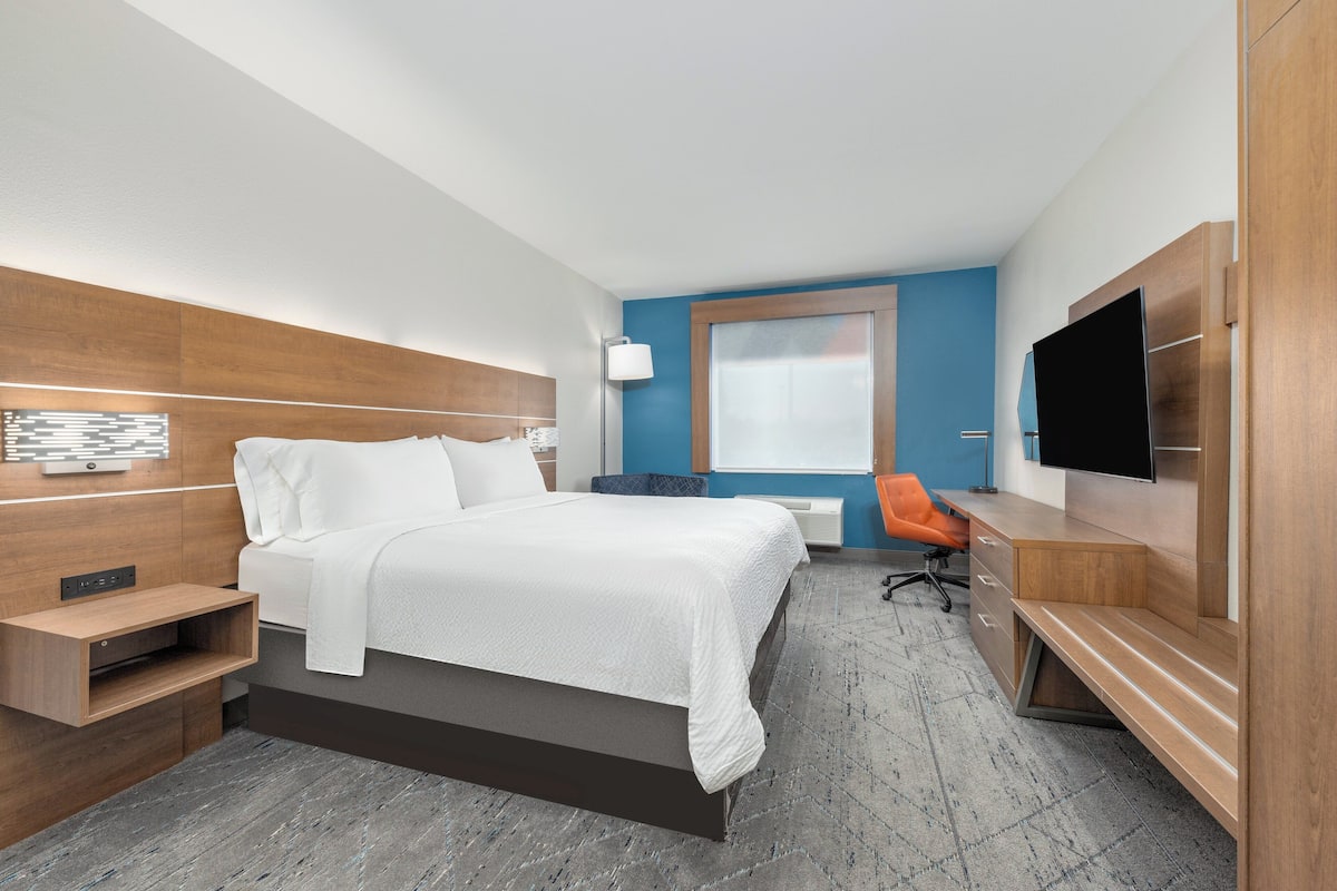 A modern hotel room with a large bed, a desk, a chair, a TV, and a window with blinds.