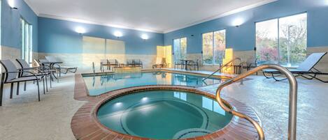 A large indoor swimming pool with a brick border, surrounded by lounge chairs and tables, and large windows offering views of trees.