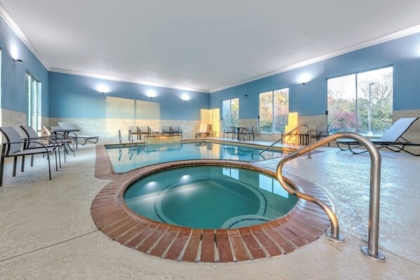 A large indoor swimming pool with a brick border, surrounded by lounge chairs and tables, and large windows offering views of trees.