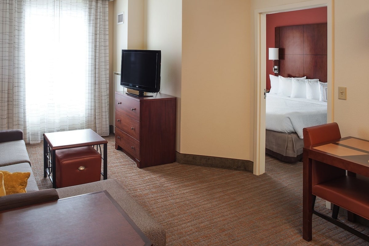 Quarto de hotel com sofá, mesa pequena, TV, escrivaninha com cadeiras, cama e banheiro visível por uma porta aberta.