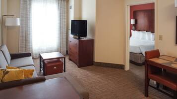 A hotel room with a sofa, a small table, a TV, a desk with chairs, a bed, and a bathroom visible through an open door.