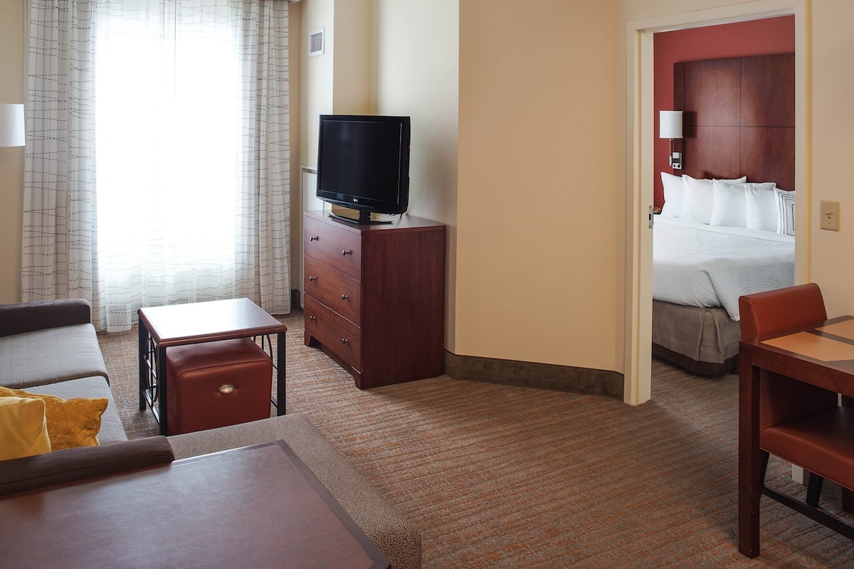Quarto de hotel com uma cama, televisão em uma cômoda de madeira, um sofá, uma mesa pequena e piso coberto por carpete.