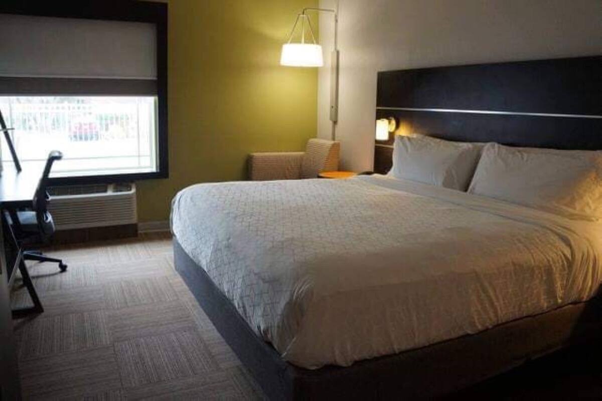 A hotel room with a large bed, a desk with a chair, a lamp, and a window with blinds.