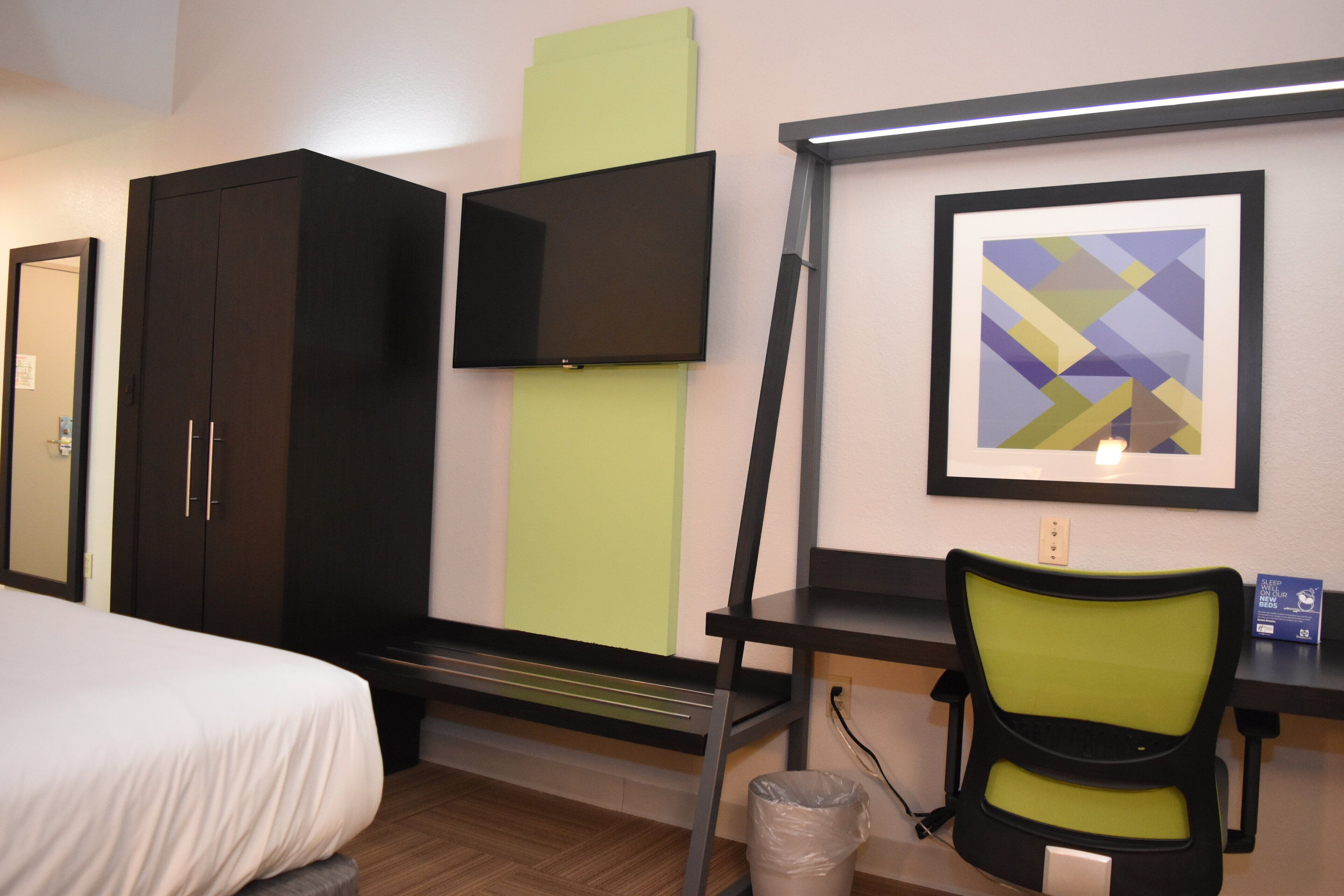 Habitación de hotel con cama, escritorio con silla, televisor, un cuadro enmarcado y un cubo de basura.