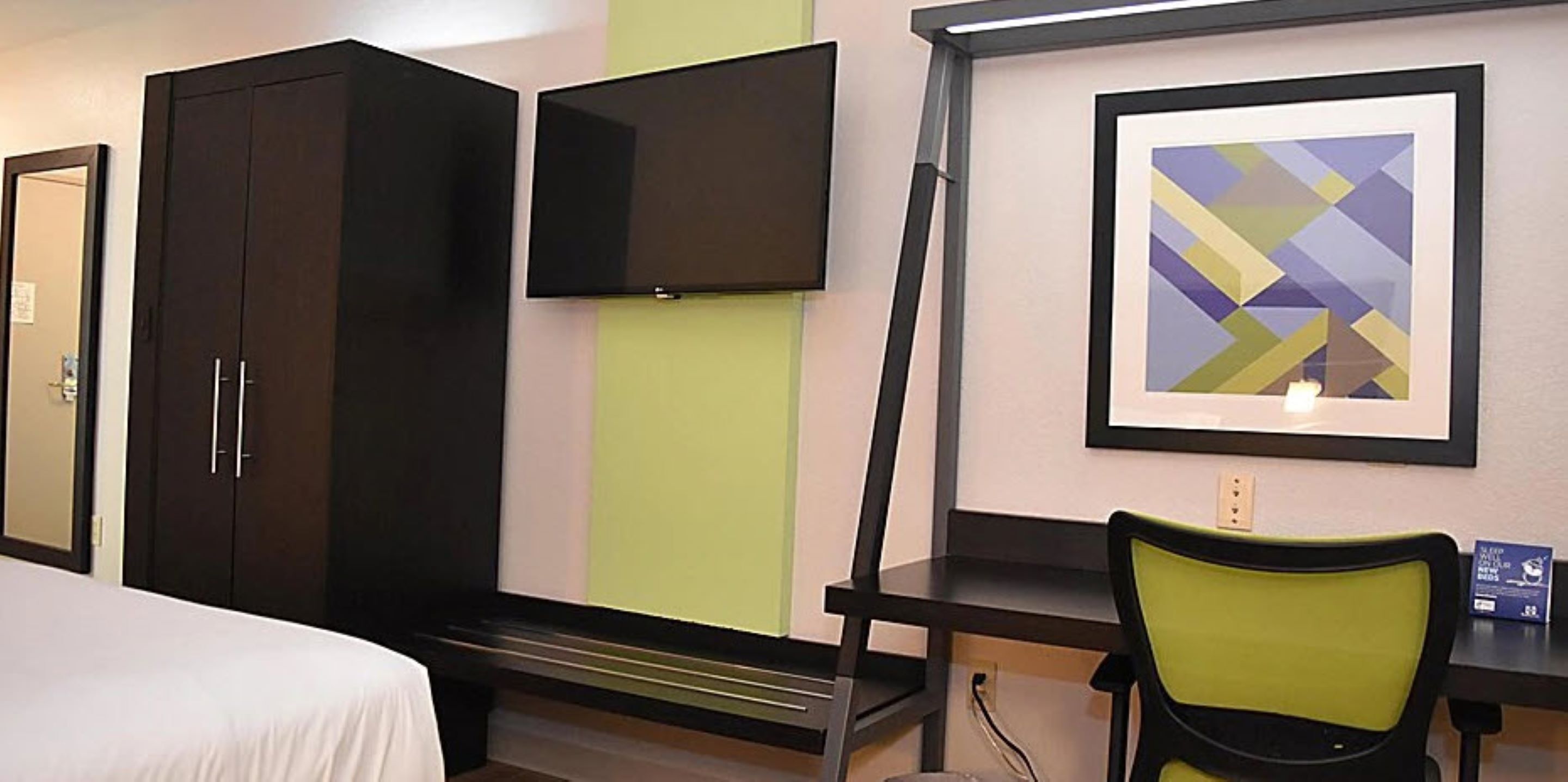 Habitación de hotel con cama, armario, televisor de pantalla plana, cuadro abstracto enmarco y escritorio con silla.