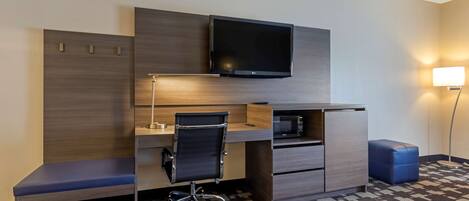 A hotel room with a wooden desk, a black office chair, a flat-screen TV mounted above the desk, a microwave, a small blue ottoman, and a floor lamp.