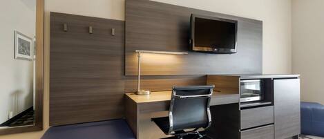 Habitación de hotel compacta con escritorio, silla, TV empotrada, microondas y un pequeño rincón de descanso.
