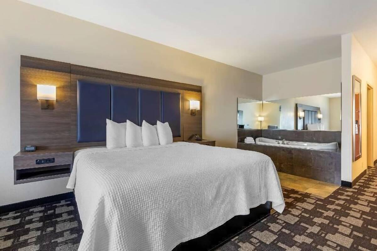 A hotel room with a large bed, a built-in headboard, bedside lamps, a mirror, and a bathroom with a bathtub.