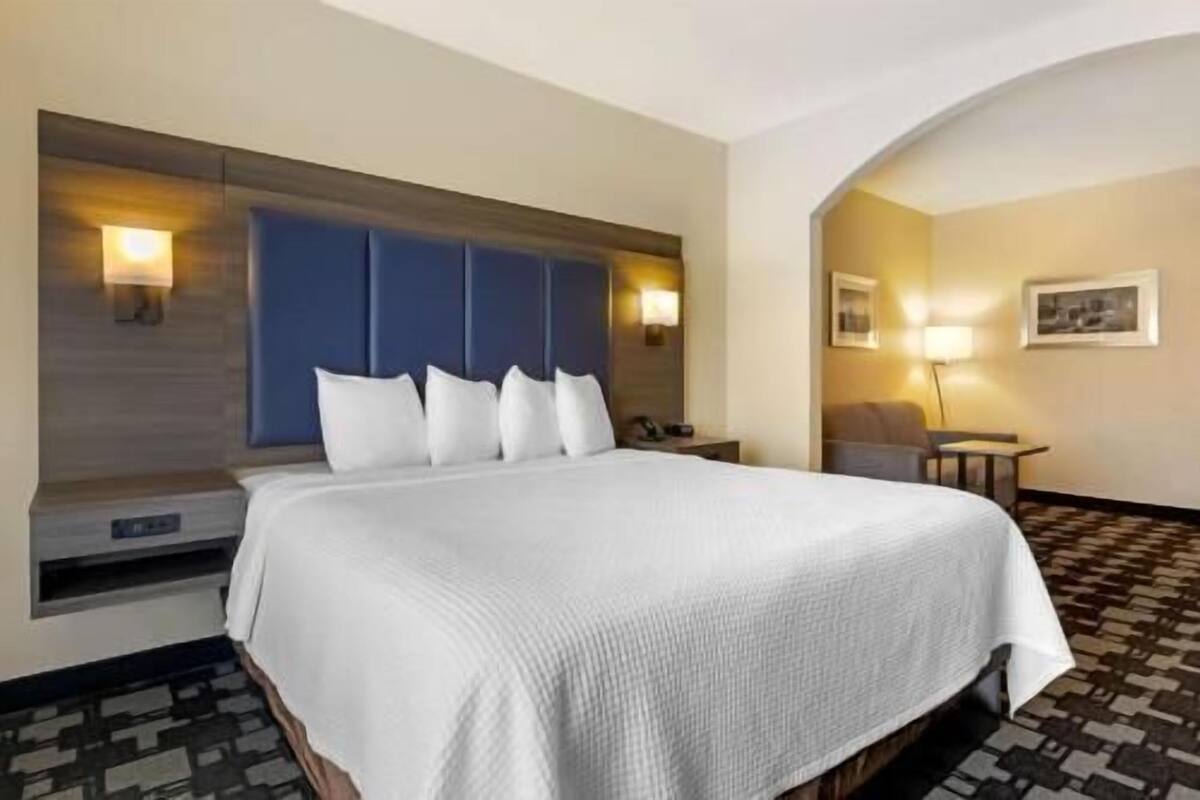 A hotel room with a large bed, a headboard with wall-mounted lights, a bedside table, a chair, and a small table with a lamp.