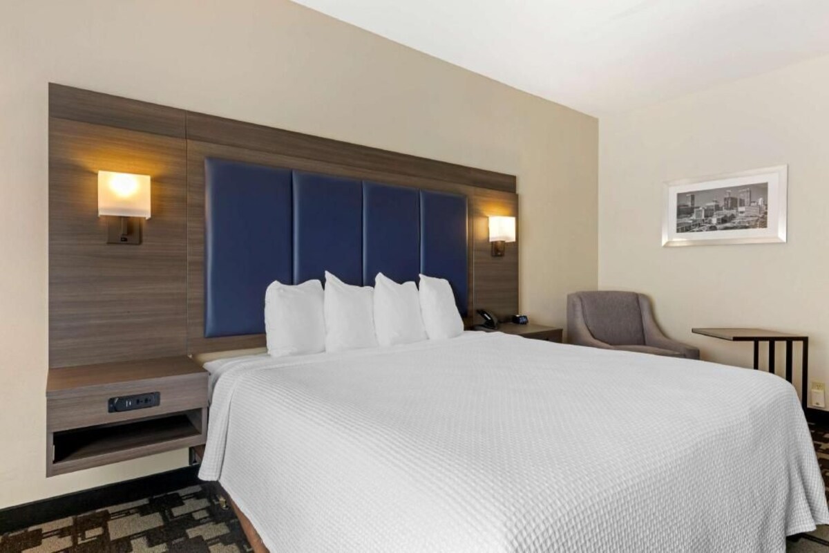 A hotel room with a large bed, a headboard with built-in lighting, a chair, a small table, and a framed picture on the wall.