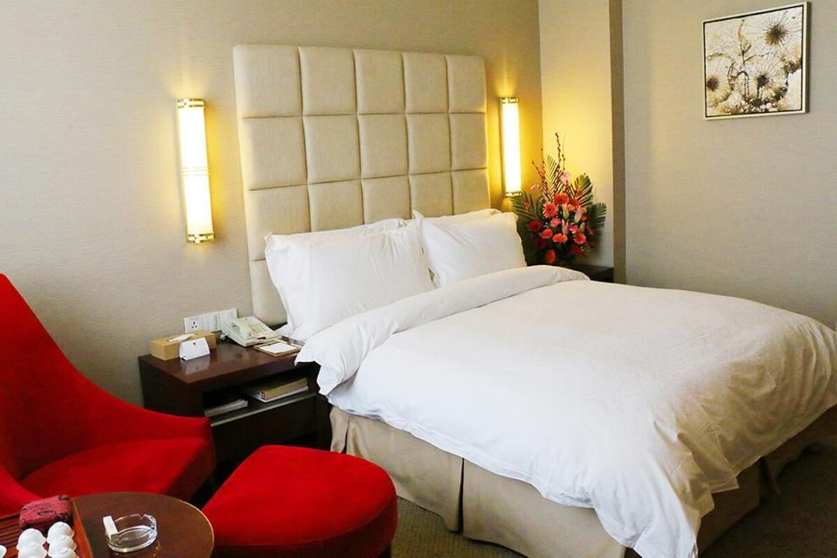 Una habitación de hotel con una cama, un sillón rojo, una mesita con un teléfono y un jarrón de flores.