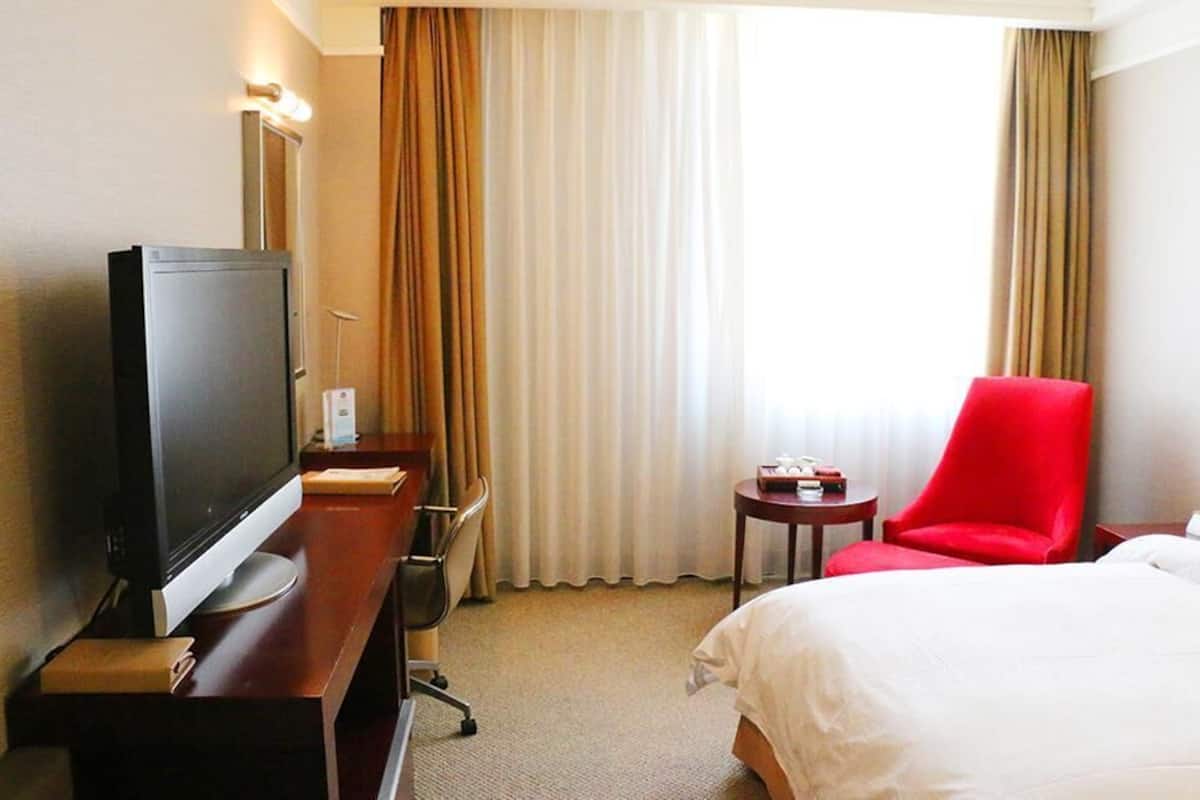 Une chambre d’hôtel avec un lit, un bureau avec une télévision, une chaise rouge et une fenêtre avec des rideaux.