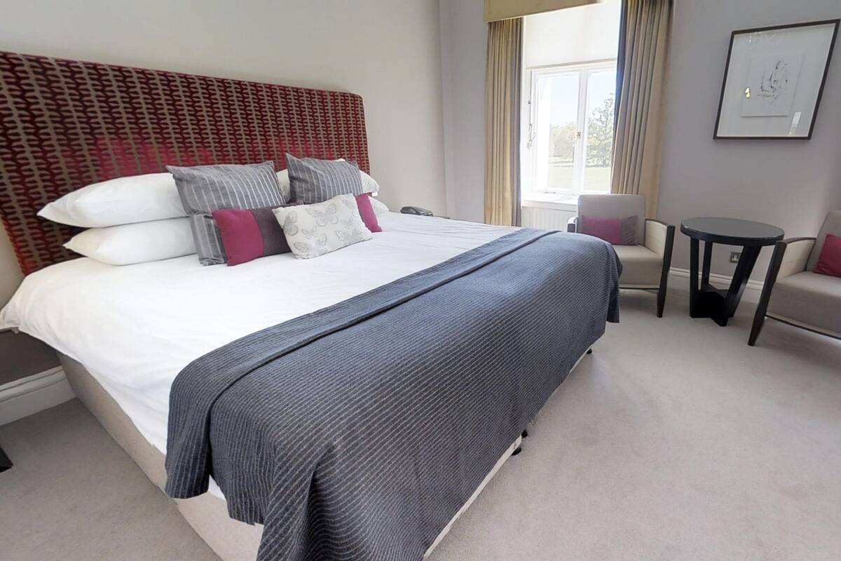 A hotel room with a large bed, a small round table, two armchairs, and a window with curtains.