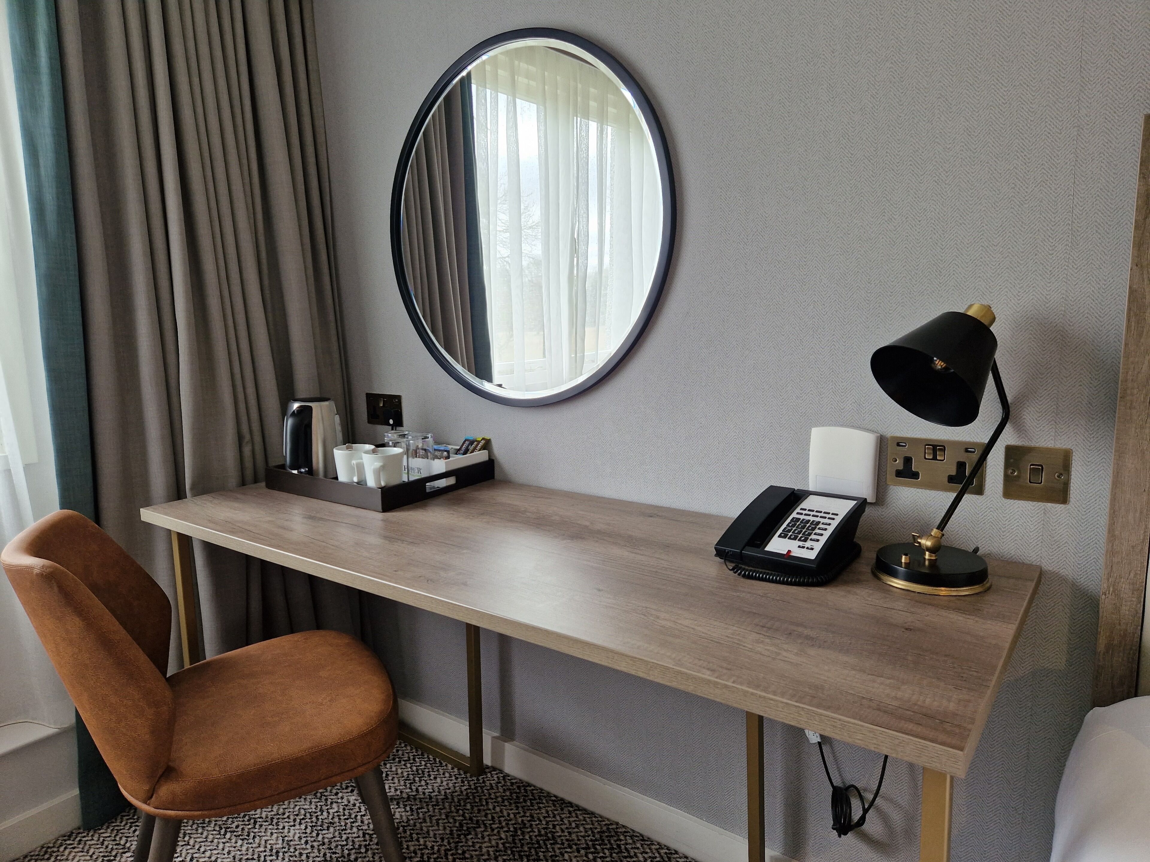 Habitación de hotel con un escritorio de madera, una silla marrón, un espejo redondo, una lámpara de escritorio negra, un teléfono y una bandeja con artículos de aseo.