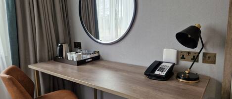 Habitación de hotel con un escritorio de madera, una silla marrón, un espejo redondo, una lámpara de escritorio negra, un teléfono y una bandeja con artículos de aseo.