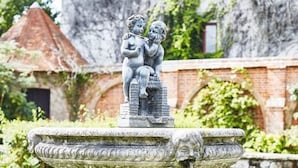 Una fuente de piedra con una escultura de dos figuras, rodeada de follaje y un muro de ladrillo.