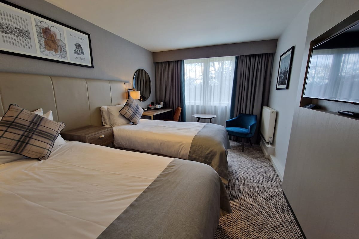 A hotel room with two beds, a desk, a chair, a TV, and a window with curtains.