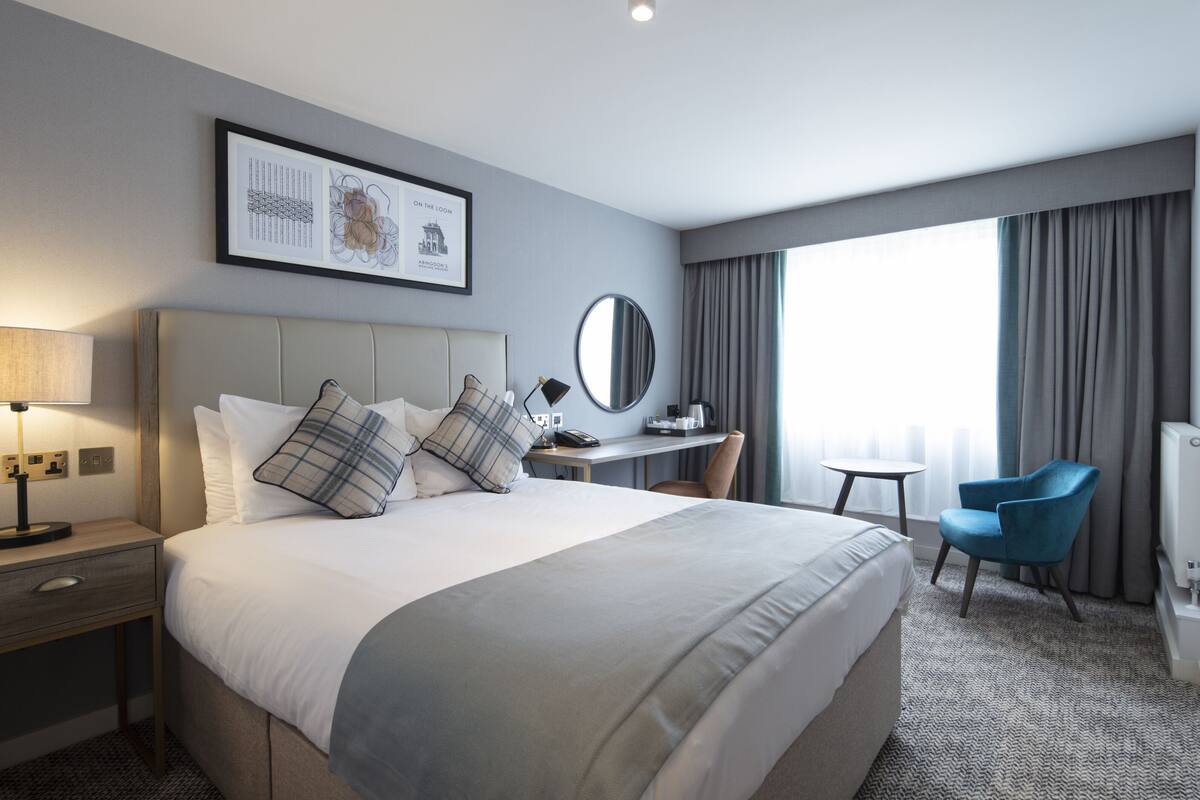 A hotel room with a large bed, a desk with a chair, a television, and a window with curtains.