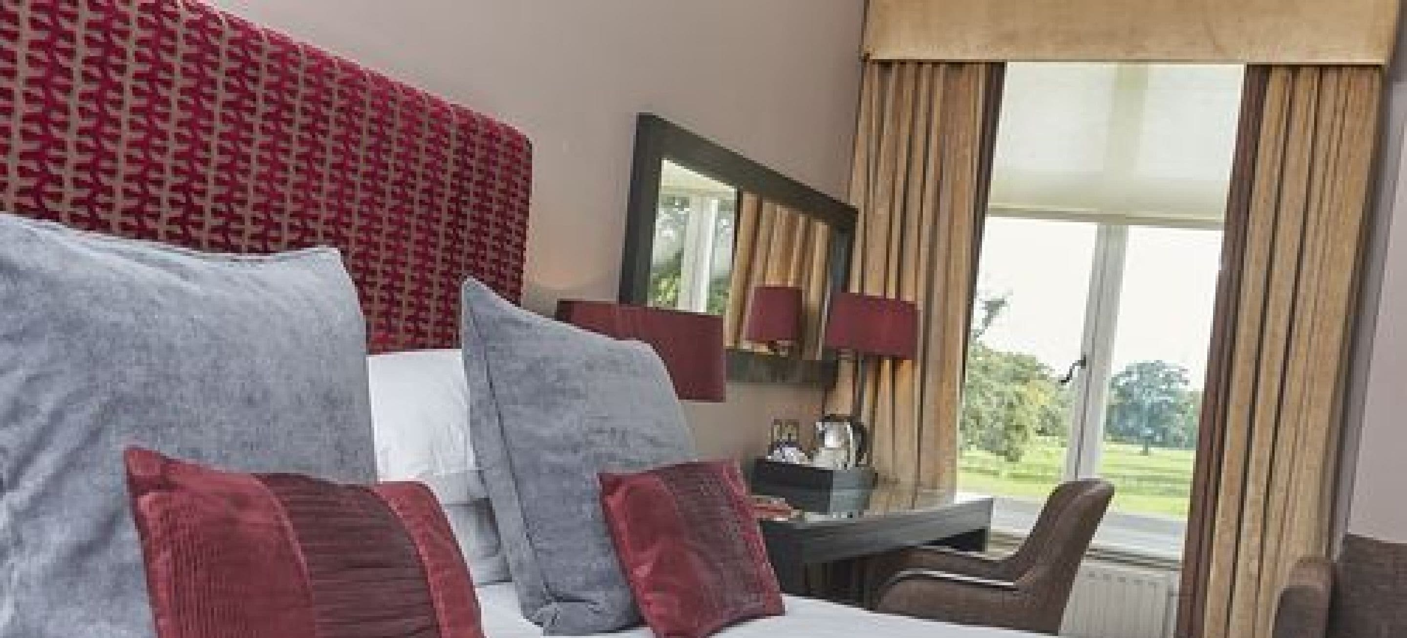 A hotel room with a bed, a desk, and a chair. There is a window with curtains and a view of greenery outside.