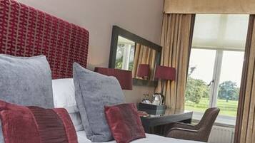 A hotel room with a bed, a desk, and a chair. There is a window with curtains and a view of greenery outside.