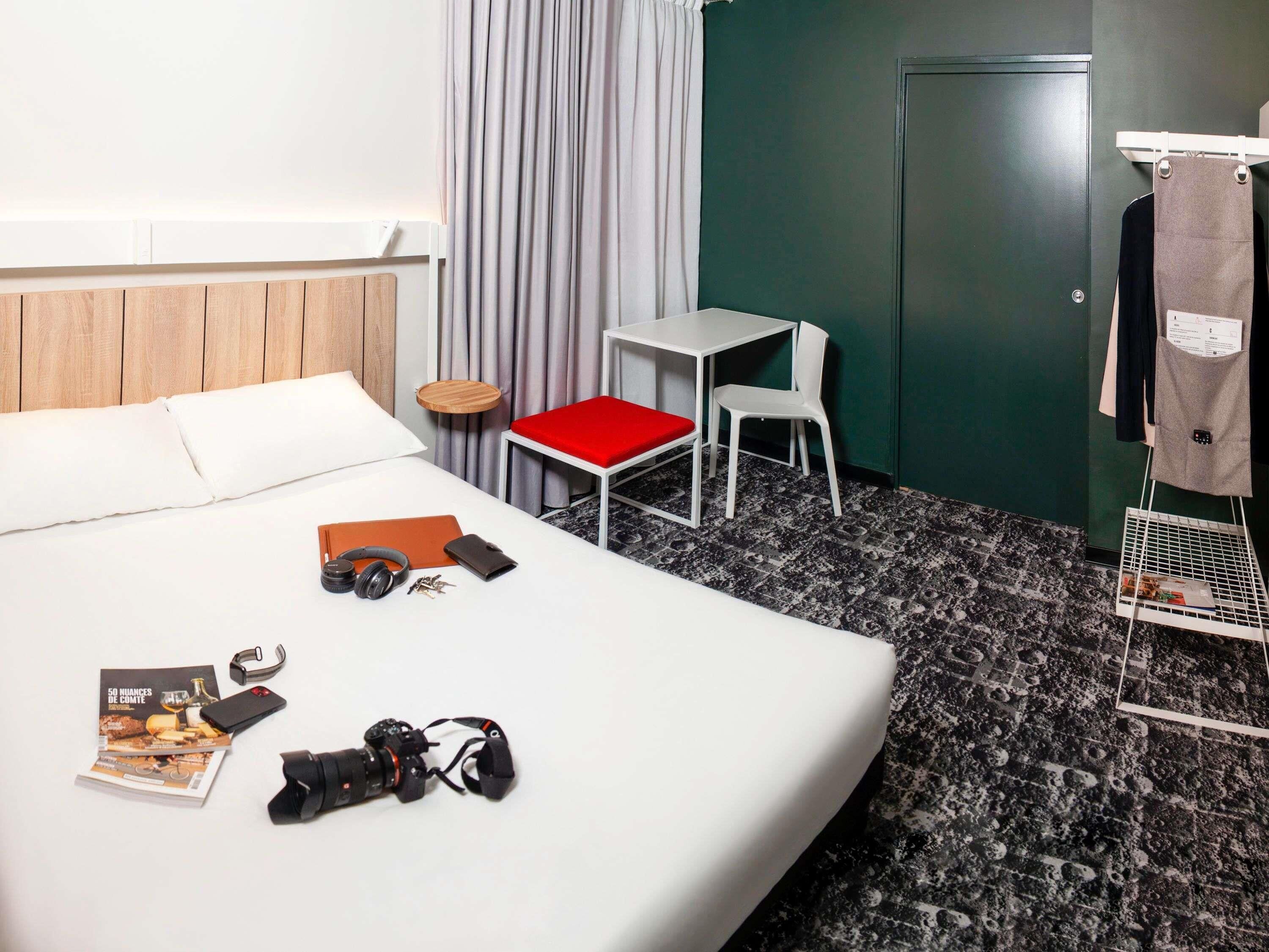Una camera d'albergo con un letto, un tavolino con una sedia, un cuscino rosso, un appendiabiti con una giacca e una macchina fotografica sul letto.