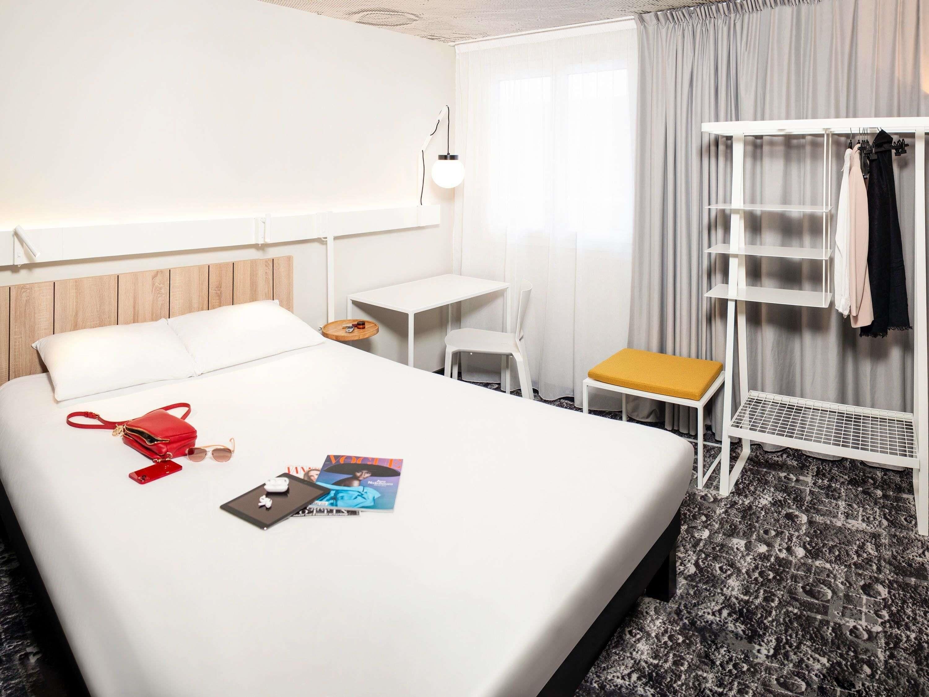 A hotel room with a bed, a desk, a chair, a small table, a shelf, and a window with curtains.