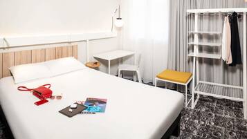 A hotel room with a bed, a desk, a chair, a small table, a shelf, and a window with curtains.