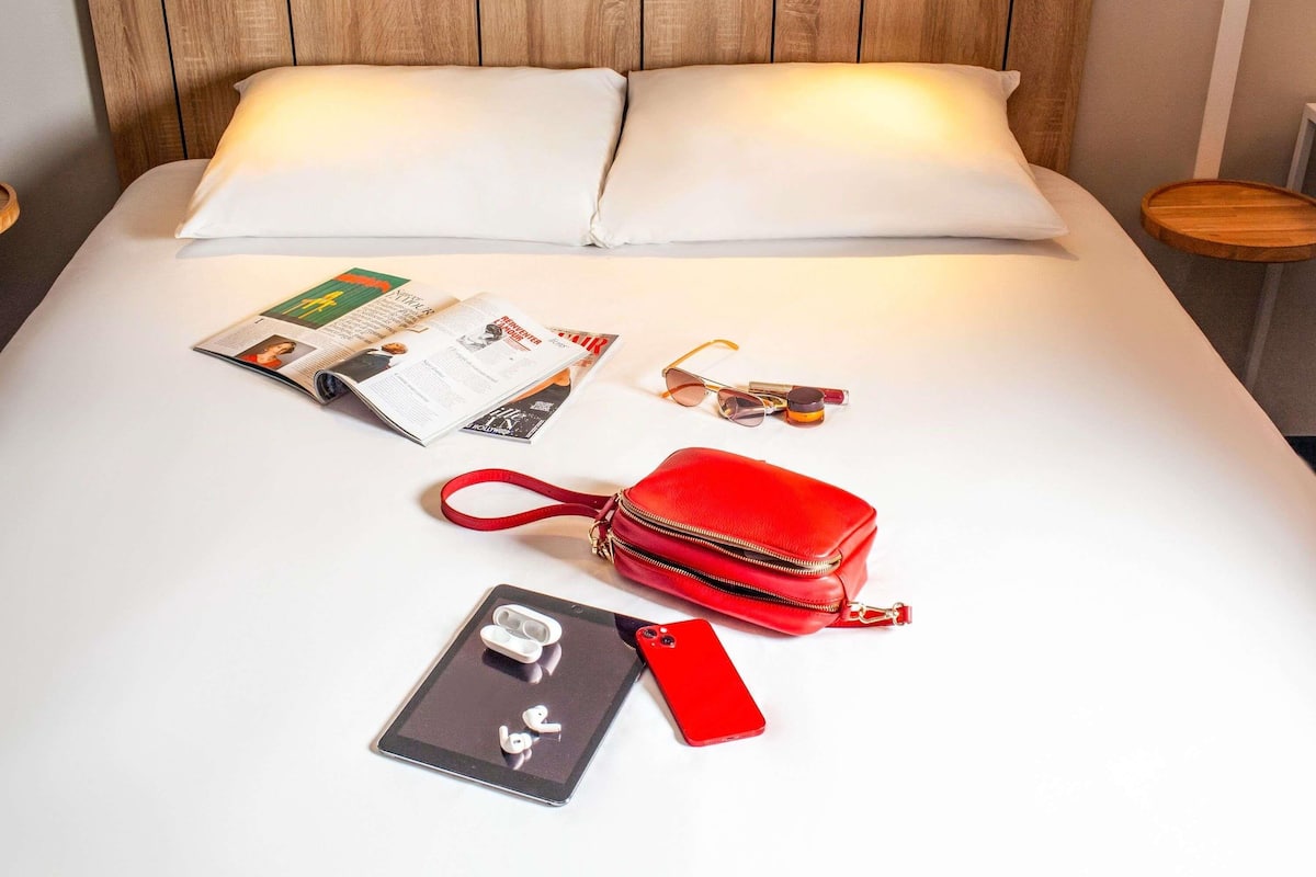 Ein ordentlich bezogenes Bett mit einem Holz-Kopfteil, eine rote Handtasche, eine Sonnenbrille, eine Zeitschrift und ein Smartphone mit Kopfhörern.