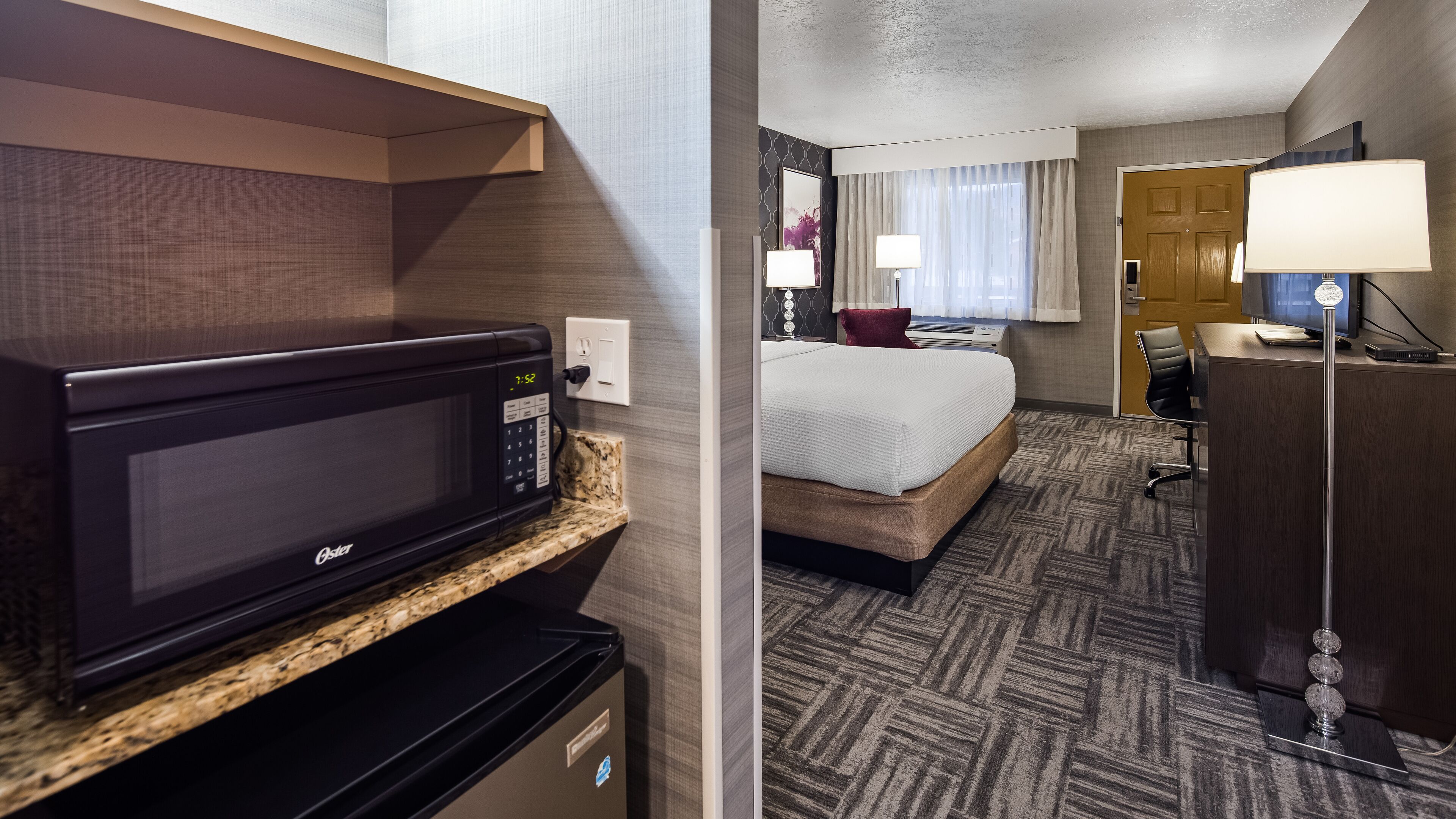 Una habitación de hotel con kitchenette, una cama, un escritorio y una lámpara.