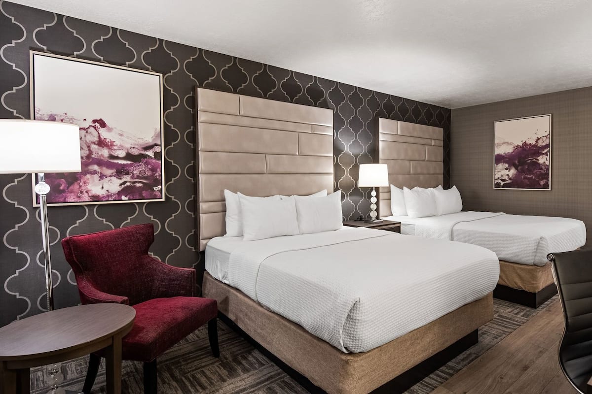 Habitación de hotel con dos camas, un sillón rojo, una mesita redonda y obras de arte en las paredes.