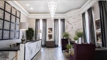 A spacious hotel lobby with a marble reception desk, a chandelier, and a comfortable seating area with a sofa and armchairs.