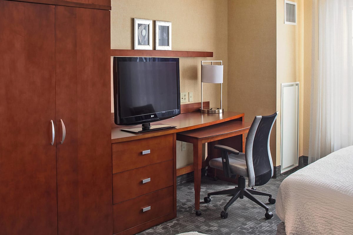 A hotel room with a wooden desk, a television, a chair, a bed, and a large wardrobe.