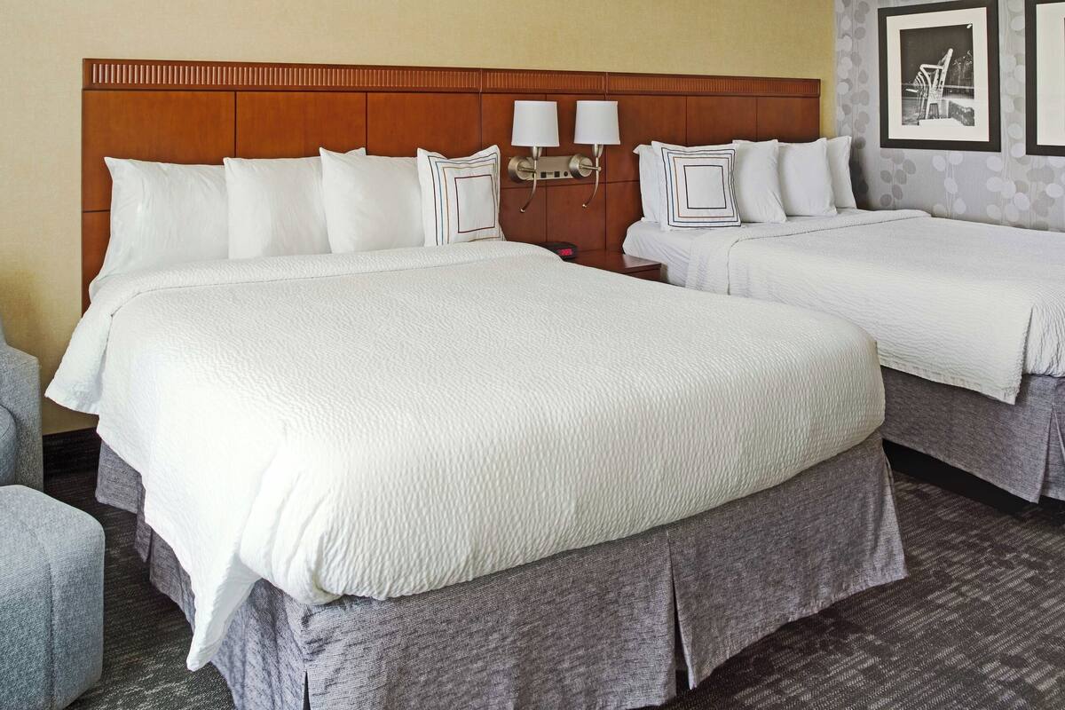 A hotel room with two beds, a wooden headboard, white bedding, and a nightstand with a lamp.