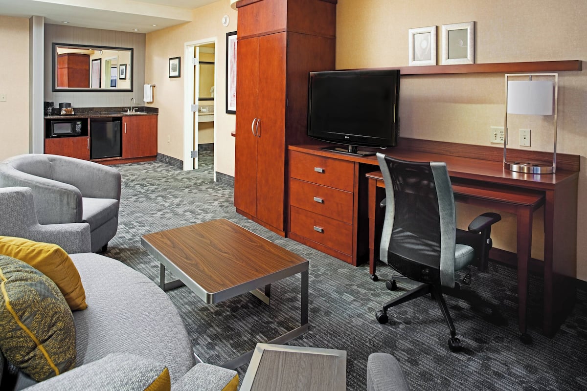 A hotel room with a sofa, a desk with a computer, a television, a small kitchenette, and a mirror.