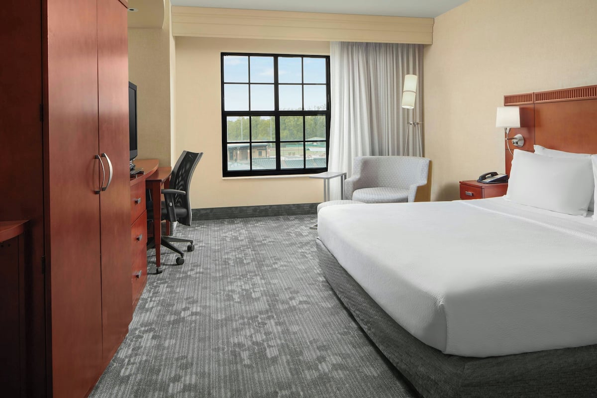 A hotel room with a large bed, a desk with a chair, a wardrobe, a window with curtains, and a nightstand with a lamp.