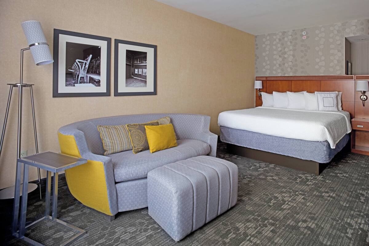A hotel room with a bed, a sofa, an ottoman, a floor lamp, and two framed pictures on the wall.