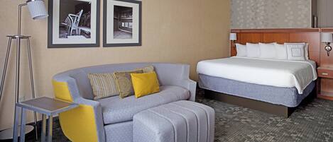 A hotel room with a bed, a sofa, an ottoman, a floor lamp, and two framed pictures on the wall.