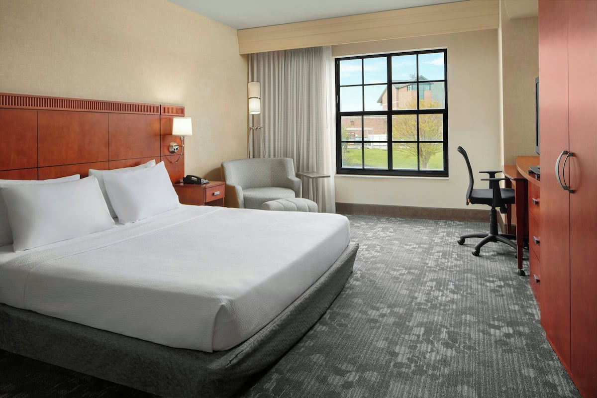 A hotel room with a large bed, a desk, a chair, a lamp, and a window with a view of buildings.