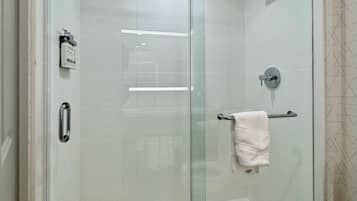 Cabina de ducha con paredes de vidrio, ducha de mano, barra para toallas y un inodoro visible en la habitación contigua.