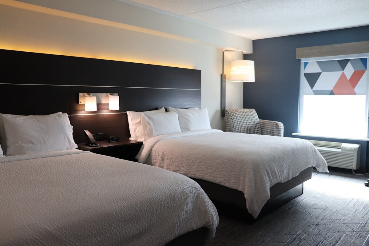 A hotel room with two beds, a nightstand with a lamp, a chair, and a large window with a geometric view.