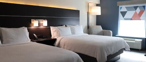 Ett hotellrum med två sängar, ett nattduksbord med en lampa, en stol och ett stort fönster med utsikt över ett område med geometriska former.