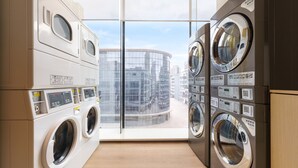 A laundry room with multiple washing machines and dryers, a control panel, and a view of a modern building through a glass wall. - Holiday Inn Express Dalian City Centre by IHG (Dalian)