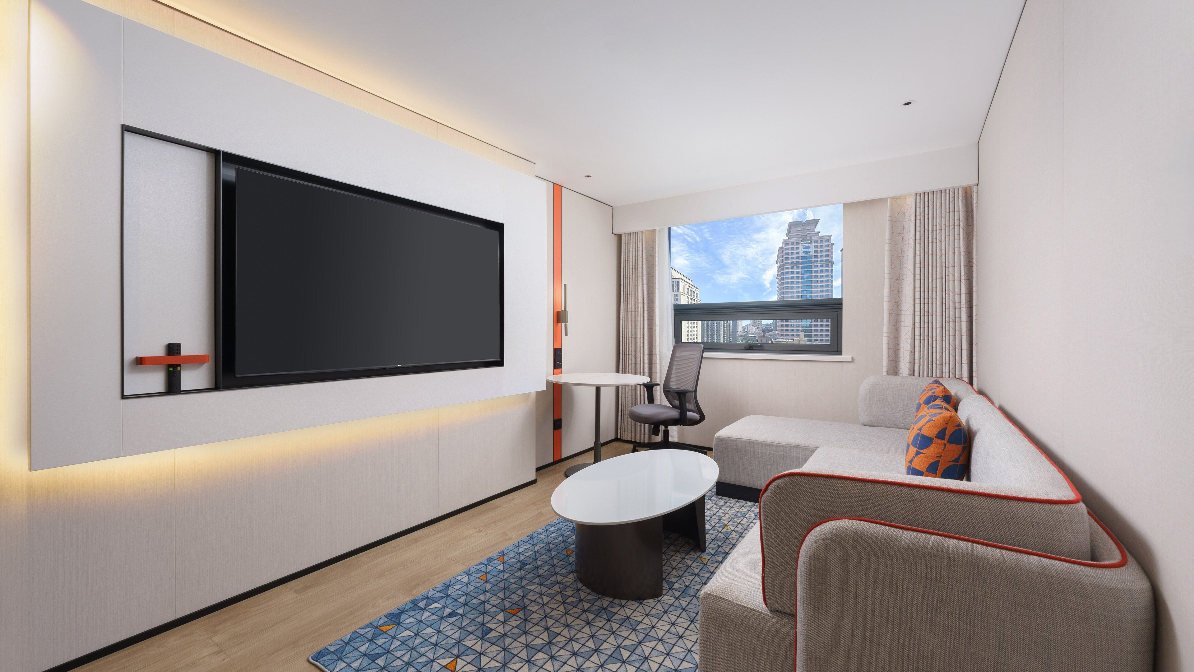 Une chambre d’hôtel moderne dotée d’un grand téléviseur à écran plat, d’un canapé, d’une petite table ronde et d’un tapis à motifs.