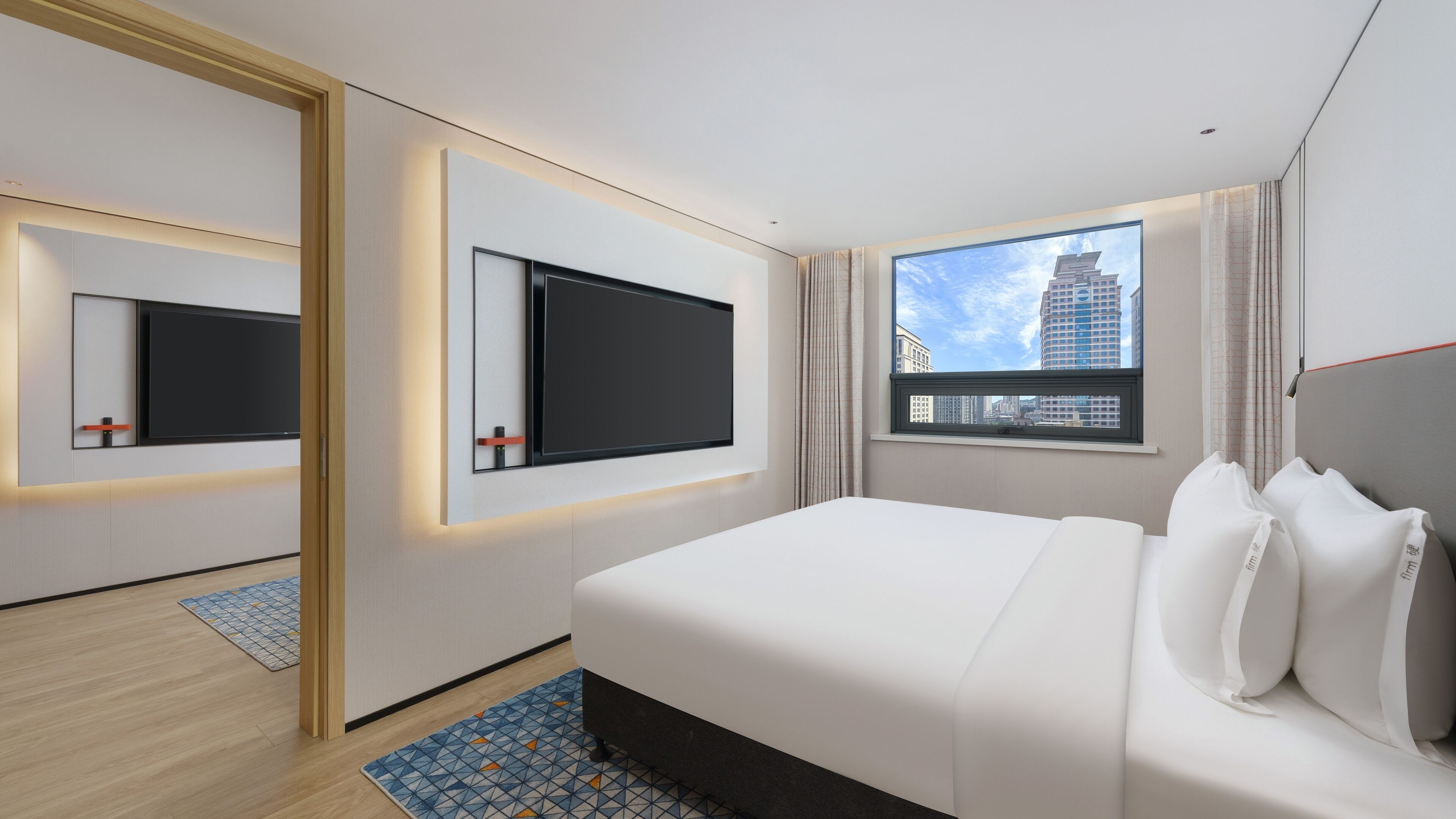 Una habitación de hotel moderna con una cama grande, dos televisores fijos en la pared, un ventanal con vista a la ciudad y una alfombra estampada.