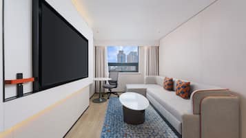 A hotel room with a large flat-screen TV, a sofa, a small table, and a view of the cityscape through the window.