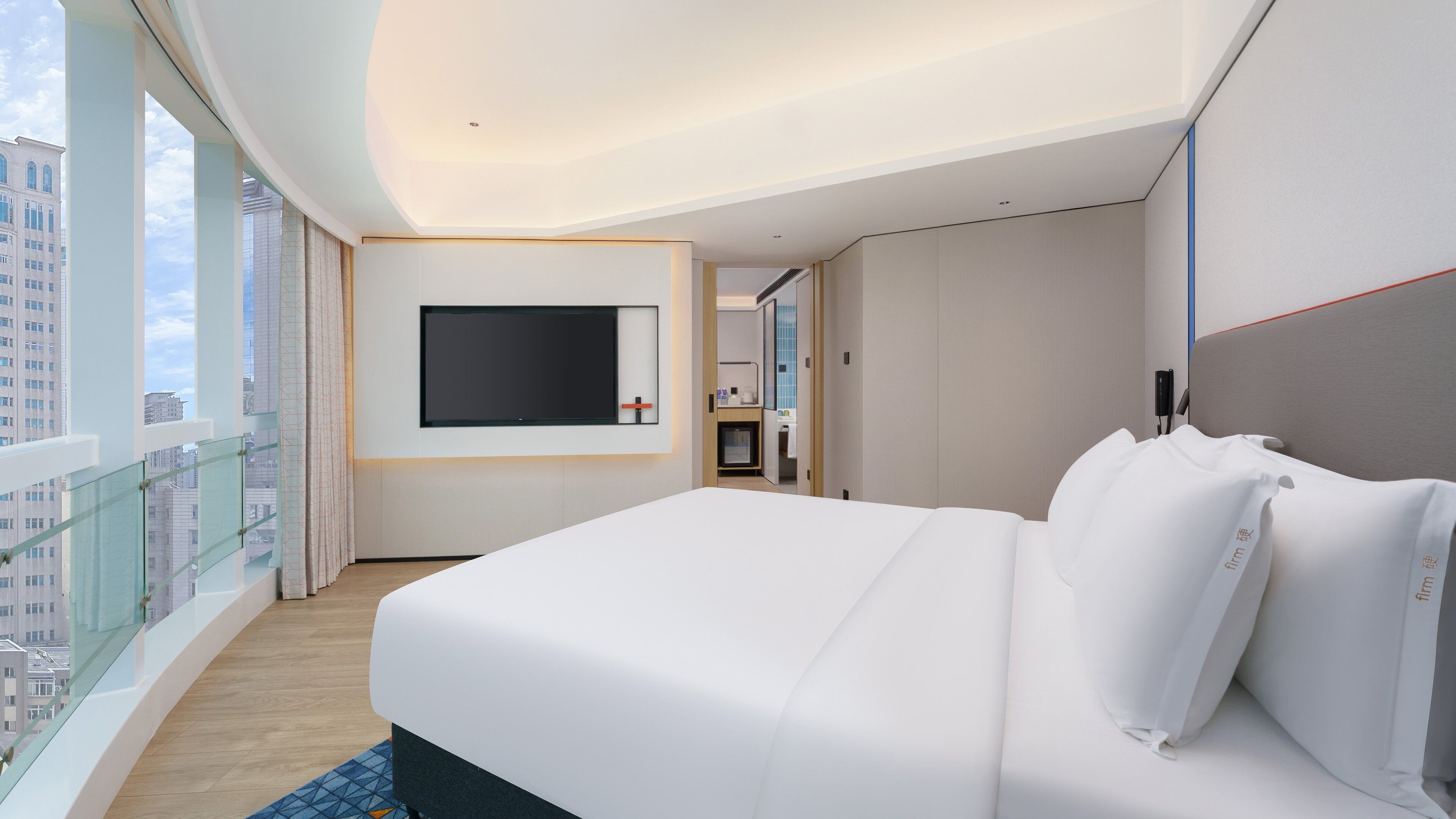 Ein Hotelzimmer mit einem großen Bett, einem wandmontierten Flachbildfernseher und Blick auf die Stadt durch große Fenster.
