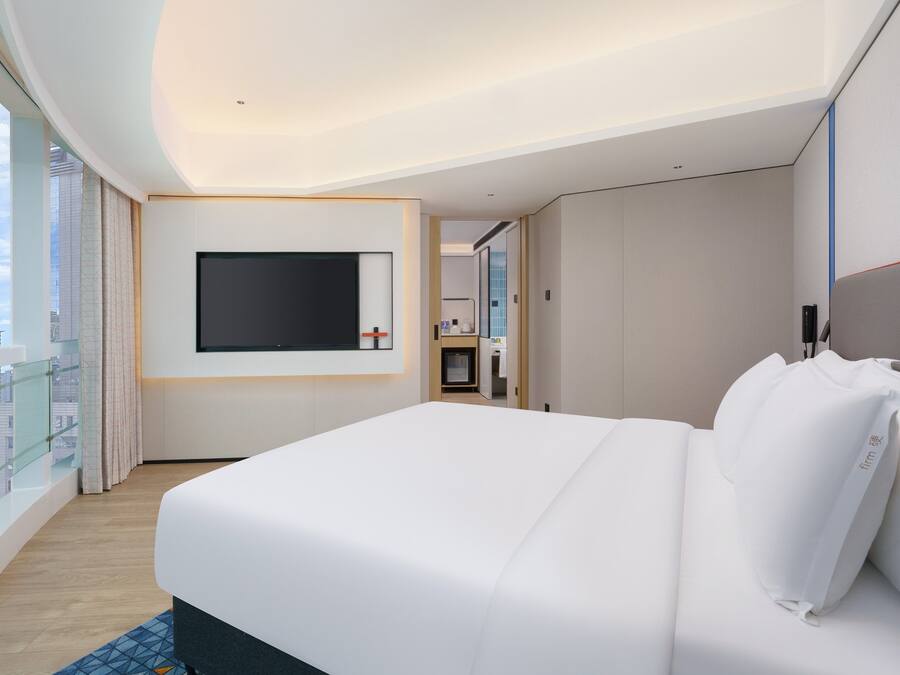 Habitación de hotel con una cama grande, un televisor de pantalla plana en la pared y vistas de la ciudad a través de amplios ventanales.