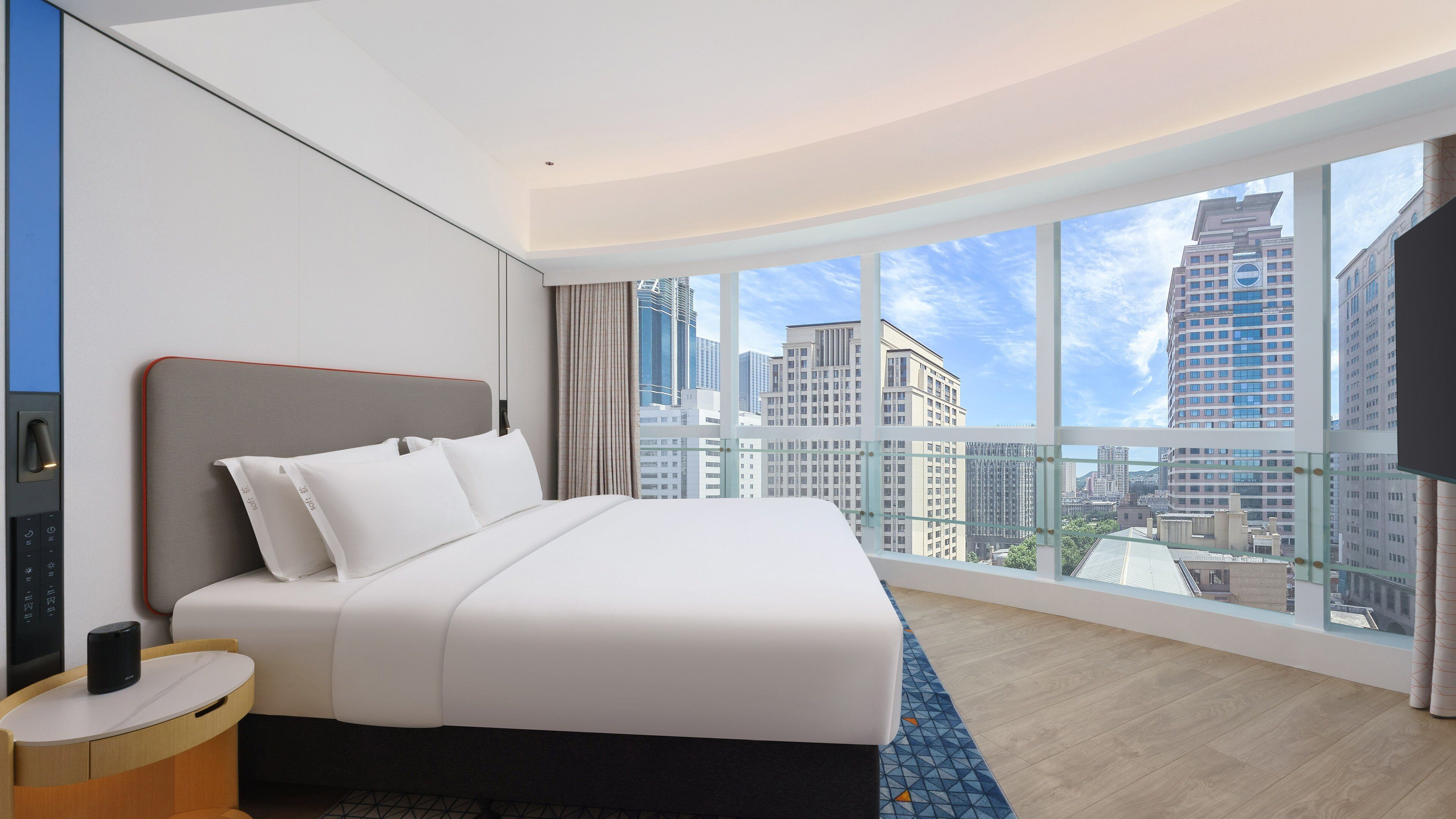 A modern hotel room with a large bed, a bedside table, a TV, and a city view.