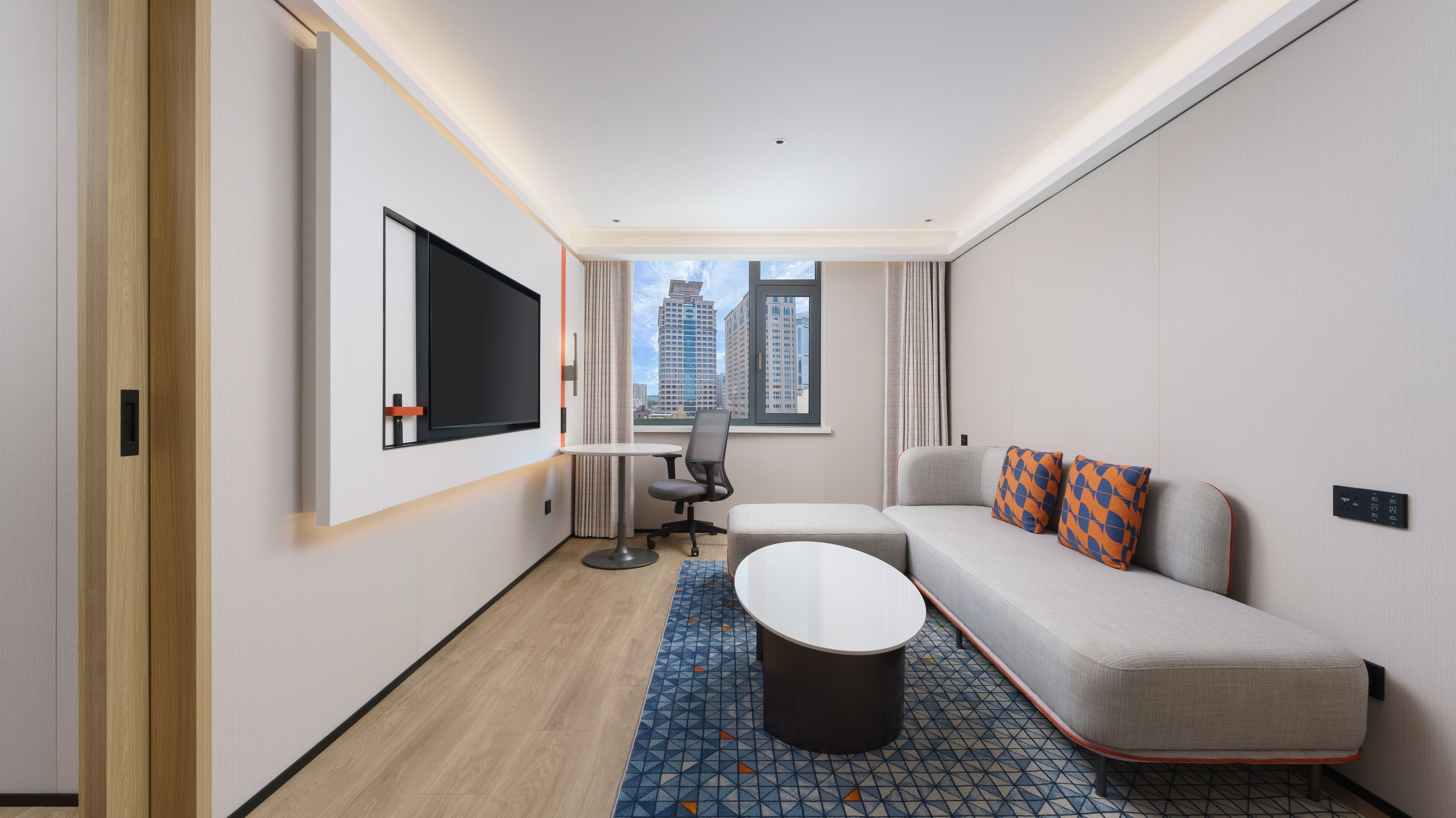Una habitación de hotel moderna con un sofá, una pequeña mesa, un televisor montado en la pared y un ventanal con vistas a la ciudad.