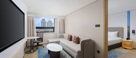 A modern hotel room with a sofa, a small table, a bed, a desk, and a large window offering a city view.